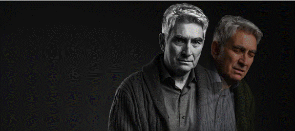 The image shows a person with grey hair and a black shirt, wearing a dark jacket, standing against a dark background.
AI-generated content may be incorrect.